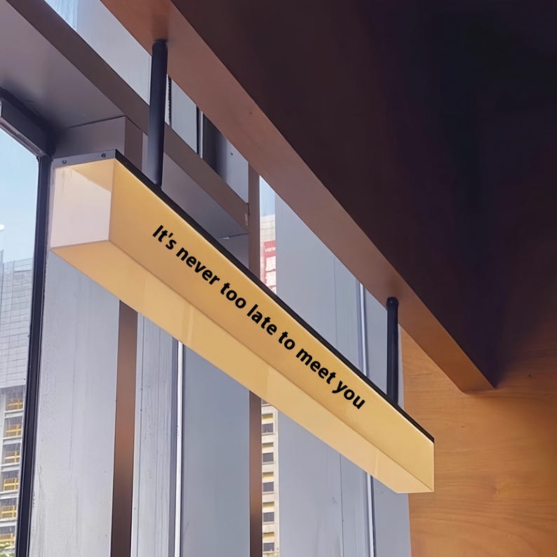 May include: A rectangular, illuminated sign with the text "It's never too late to meet you" in black. The sign is suspended from the ceiling with black rods. The sign has a yellow glow, and is near a window.
