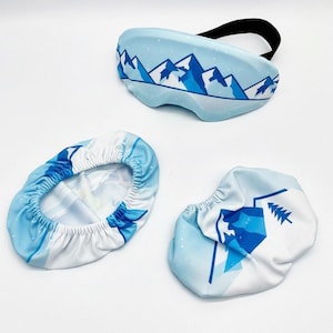 May include: A light blue ski goggle cover and two matching drawstring covers. The goggle cover has a black strap and a mountain range design in shades of blue. The drawstring covers have a similar mountain and tree design.
