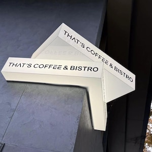 May include: A white, angular sign for a coffee shop and bistro. The sign is mounted on a gray wall and features the text "THAT'S COFFEE & BISTRO" in black, bold lettering. The sign has a modern, geometric design.