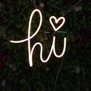 Pode incluir: Uma placa de néon branca com a palavra "hi" e um coração no meio do "i". A placa está pendurada em uma parede verde.