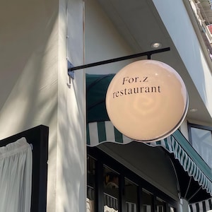 Peut inclure: Un enseigne de restaurant sphérique et éclairée, de couleur crème, avec le texte "For.z restaurant" en noir. Le panneau est monté sur un bras métallique noir. Un auvent à rayures vertes et blanches est visible en dessous.