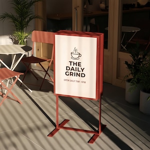 Puede incluir: Un letrero metálico rojo con el texto "THE DAILY GRIND" y un logotipo de taza de café. El letrero está sobre un soporte y se coloca fuera de un café. El letrero también incluye el horario: ABIERTO DIARIAMENTE 7AM - 6PM.