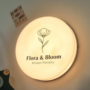 May include: A round, illuminated sign with the text "Flora & Bloom Artisan Floristry" and a floral illustration. The sign is white with a soft glow, and a small decorative card is visible in the upper left corner.