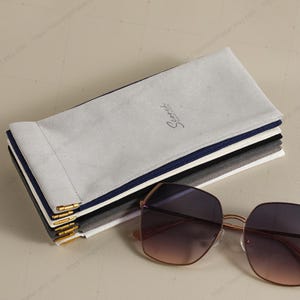 May include: A pair of oversized, geometric sunglasses with a gradient lens, next to a stack of gray, black, and navy blue eyeglass cases. The cases have gold-colored metal accents and the word "Scorch" printed on the top case.