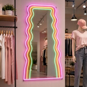 Puede incluir: Un gran espejo con bordes ondulados y un borde de luz de neón en rosa, amarillo y verde cuelga de una pared rosa. El espejo refleja percheros y un maniquí con una camiseta rosa y jeans. La tienda tiene un diseño moderno y minimalista.