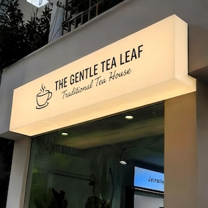 May include: Illuminated sign for "The Gentle Tea Leaf Traditional Tea House." The sign is rectangular, with a white background and black text. A stylized teacup with steam is on the left. The sign is mounted above a glass storefront.