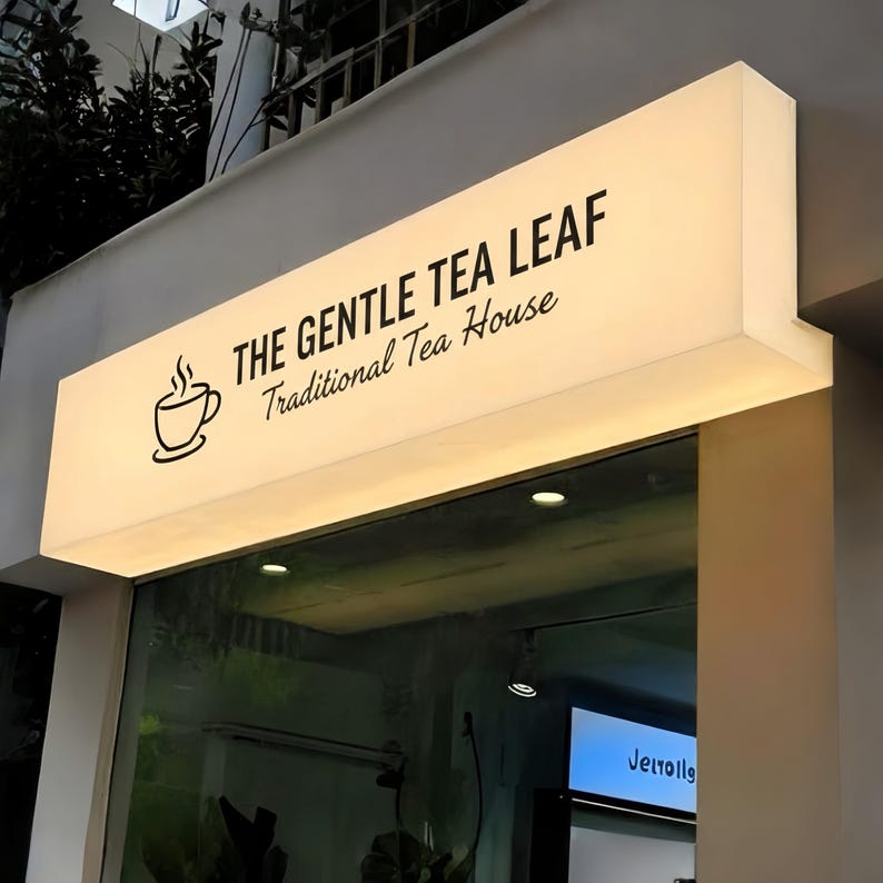 May include: Illuminated sign for "The Gentle Tea Leaf Traditional Tea House." The sign is rectangular, with a white background and black text. A stylized teacup with steam is on the left. The sign is mounted above a glass storefront.