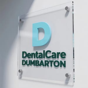 以下が含まれることがあります： 「DentalCare DUMBARTON」の文字が濃い緑色で、大きな水色の「D」が描かれた透明アクリル看板。白い壁に銀色のスペーサーで取り付けられています。