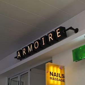 Può includere: Insegna nera illuminata che recita "ARMOIRE" in maiuscolo, montata su una parete bianca. L'insegna è composta da singoli cubi neri con le lettere ritagliate, che consentono alla luce di brillare. Una seconda insegna sottostante recita "NAILS MASSAGE".