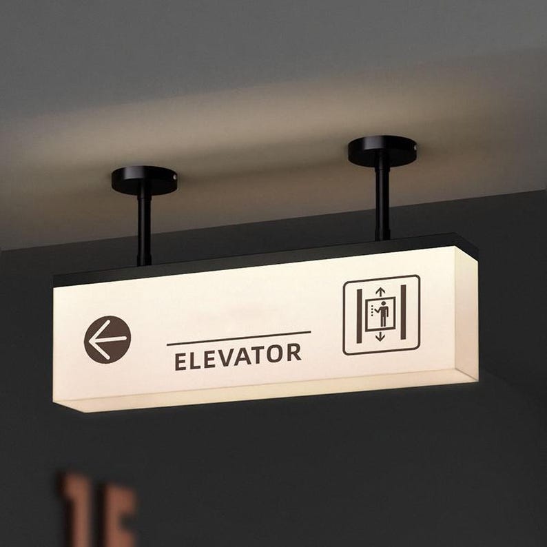 May include: A rectangular, illuminated elevator sign with black text and symbols. The sign reads "ELEVATOR" with an arrow pointing left and an elevator symbol on the right. The sign is suspended from the ceiling by two black rods.