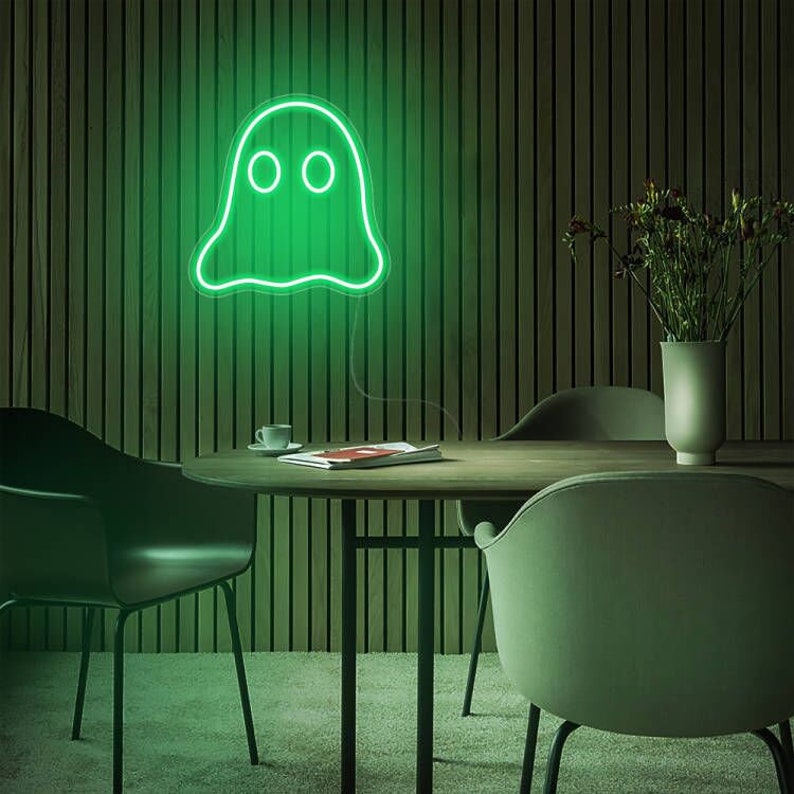 May include: A green neon ghost sign is displayed on a wooden wall. The ghost has two round eyes and a wavy outline. A round wooden table with chairs, a vase with flowers, and a coffee cup are in the foreground.