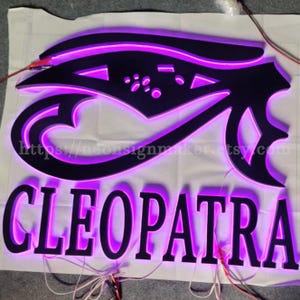 May include: A black 3D sign with the word "CLEOPATRA" in white letters. The sign is lit up with purple neon lights. The sign also features a stylised eye of Horus design.