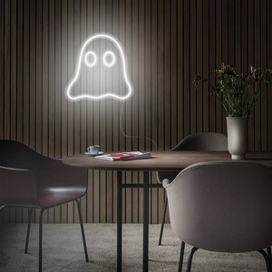 May include: A white neon ghost sign hangs on a dark wood-panelled wall. The ghost has two round eyes and a wavy bottom. The sign is above a round wooden table with chairs. A vase of flowers sits on the table.