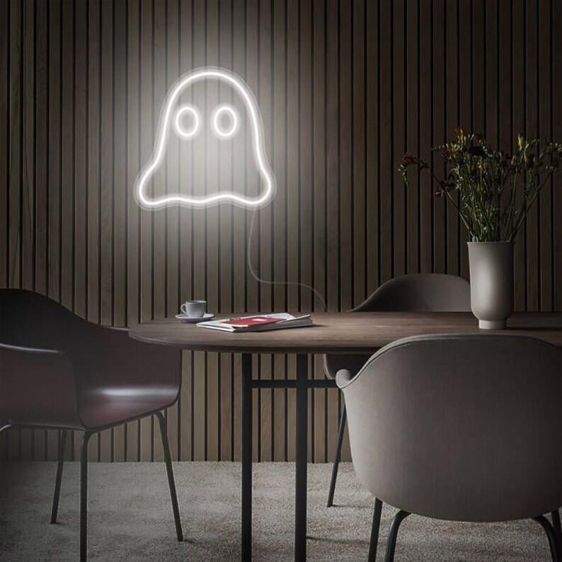 May include: A white neon ghost sign hangs on a dark wood-panelled wall. The ghost has two round eyes and a wavy bottom. The sign is above a round wooden table with chairs. A vase of flowers sits on the table.