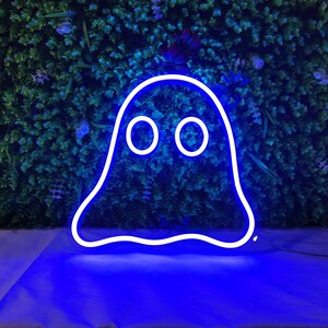 May include: A blue neon ghost sign with a white outline and two circular eyes. The neon sign is displayed against a backdrop of green foliage and purple flowers. The sign is lit up and casting a blue glow.