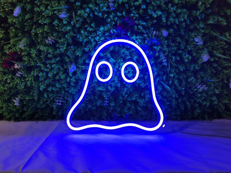 May include: A blue neon ghost sign with a white outline and two circular eyes. The neon sign is displayed against a backdrop of green foliage and purple flowers. The sign is lit up and casting a blue glow.