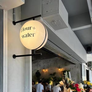 Pode incluir: Uma placa redonda, branca e iluminada, montada num suporte de metal preto fixado a uma parede. A placa apresenta o texto preto "flour + water" impresso na sua superfície. Está posicionada acima de uma entrada para um espaço iluminado.