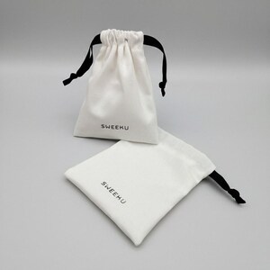 May include: Two white drawstring bags with black drawstrings. The bags are made of a soft fabric and have the word "SWEEKU" printed in black on the front. One bag is upright, and the other is lying flat.