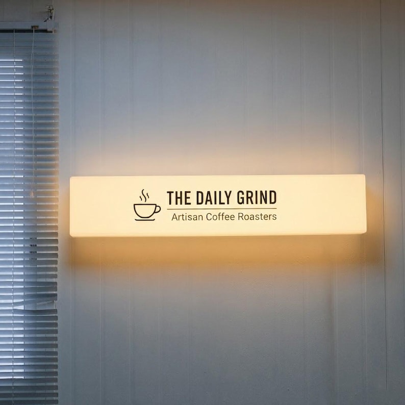 May include: A rectangular, illuminated sign with the text "THE DAILY GRIND Artisan Coffee Roasters" in black. A coffee cup icon is to the left of the text. The sign is mounted on a white wall, with a window and blinds visible on the left.