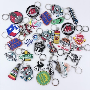 May include: A collection of various colorful rubber keychains featuring diverse graphic designs, logos, and text including Rice Fest, Zibby Towing, Pfizer 175, Gulf Coast Invitational, SJ Farm, Prime Cuts Lawn Care, and others with metal rings.
