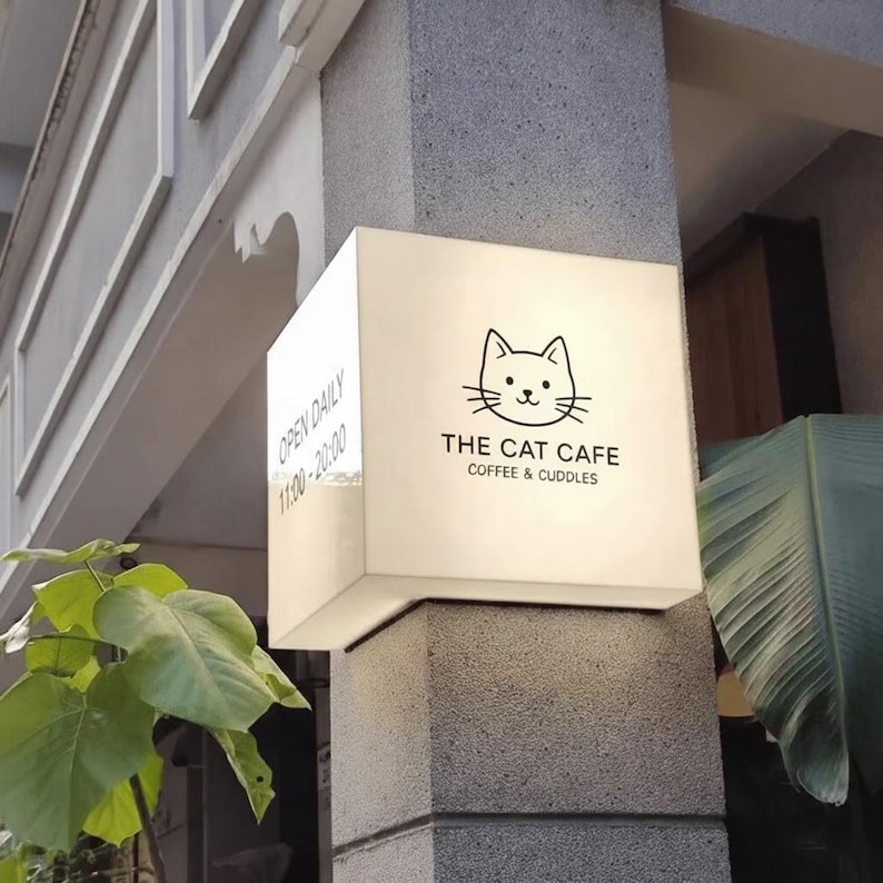 May include: A white, illuminated sign for "The Cat Cafe" with a black cat illustration and the text "Coffee & Cuddles." The sign is mounted on a gray pillar and displays the hours of operation.