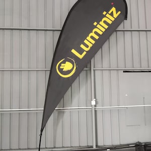 May include: A black teardrop banner with the word "Luminiz" in yellow, along with a yellow logo. The banner is mounted on a black pole and a gray base. The banner is displayed indoors.