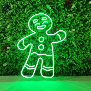May include: A vibrant green neon gingerbread man sign with a smiling face, three buttons, and striped legs. The sign is set against a backdrop of lush green foliage, creating a festive and eye-catching display. Ideal for Christmas decorations.