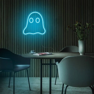 May include: A blue neon ghost sign is mounted on a wooden wall. The ghost has two round eyes and a wavy outline. The sign is above a round wooden table with chairs. A vase of flowers and a coffee cup are also on the table.