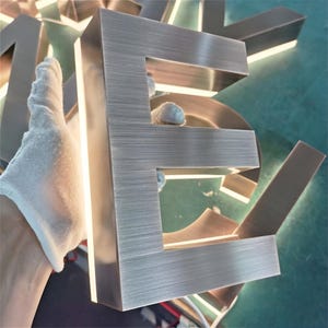 May include: A brushed metal letter E with a light source behind it, creating a halo effect. The letter is made of a durable metal and has a modern, industrial look.