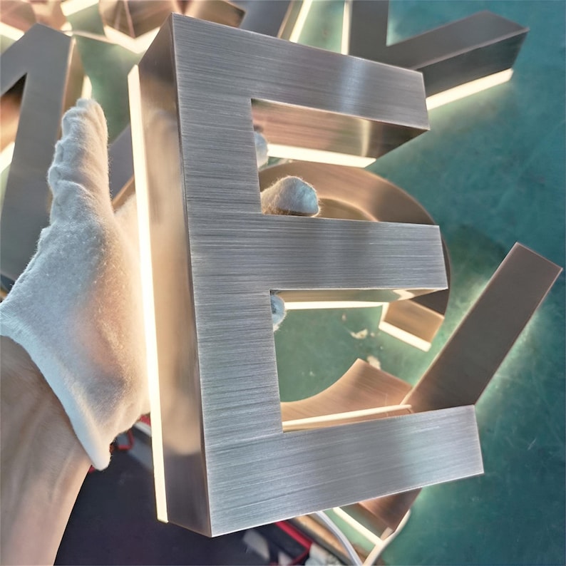 May include: A brushed metal letter E with a light source behind it, creating a halo effect. The letter is made of a durable metal and has a modern, industrial look.