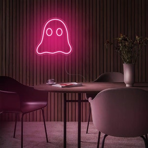 May include: A pink neon ghost sign with two round eyes is mounted on a wooden wall. The neon light is the focal point of the room, which also includes a round table, chairs, and a vase with flowers.