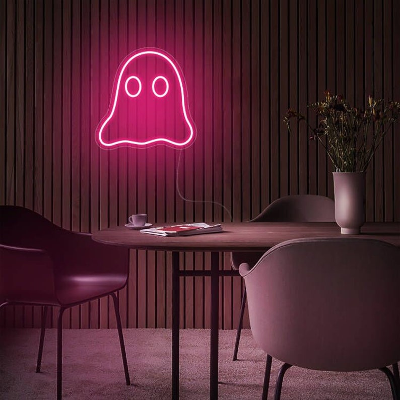 May include: A pink neon ghost sign with two round eyes is mounted on a wooden wall. The neon light is the focal point of the room, which also includes a round table, chairs, and a vase with flowers.