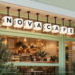 May include: A series of nine square, white, illuminated light boxes suspended from the ceiling, each displaying a single black letter to spell out NOVA CAFFEE. The signs are mounted above a storefront window with a light green frame.