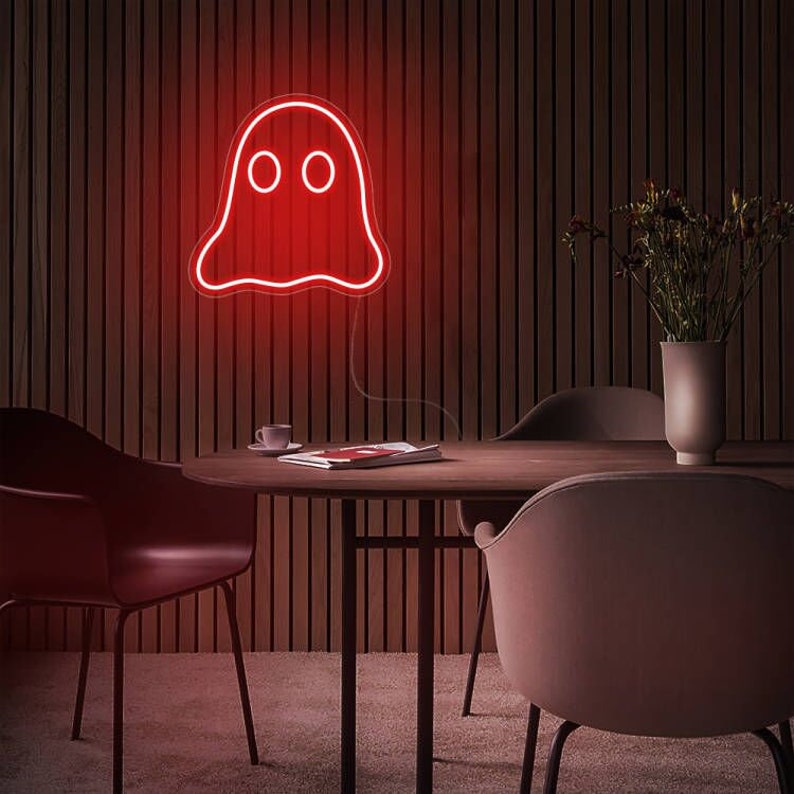 May include: A red neon ghost sign with two circular eyes, fixed to a wooden wall. The sign is lit, emitting a warm glow. The room includes a round table, chairs, and a vase of flowers, creating a comfortable atmosphere.
