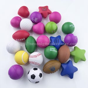 May include: A collection of colorful stress balls in various shapes, including hearts, stars, and sports balls like a golf ball, baseball, and football. The stress balls are in shades of pink, green, purple, yellow, and white, arranged on a white surface.