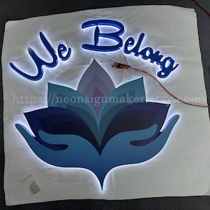 May include: A blue neon sign with the words "We Belong" in a cursive font. The sign is illuminated with white LED lights and features a blue and purple lotus flower design with hands cupping the flower.