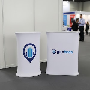 Puede incluir: Dos stands promocionales blancos. Uno tiene un logotipo azul y blanco de un edificio dentro de un pin de ubicación. El otro muestra la palabra "geotees" en azul. Ambos son cilíndricos.