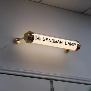 Op de afbeelding: Een aan de muur gemonteerde lamp met een heldere, cilindrische buis en goudkleurige metalen accenten. De lamp toont de tekst "SANDBAR CAMP" in zwart, samen met een afbeelding van een bergketen en een regenboog.