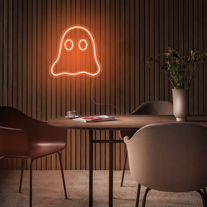 May include: Orange neon ghost sign with two round eyes, mounted on a wooden wall. The sign is illuminated, casting a warm glow. The room features a round wooden table, chairs, and a vase with flowers.