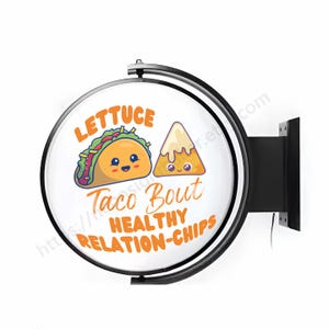 May include: A round, black-framed sign with a white background. The sign features a cartoon taco and a cartoon nacho chip with smiling faces. The text on the sign reads "Lettuce Taco Bout Healthy Relation-Chips".