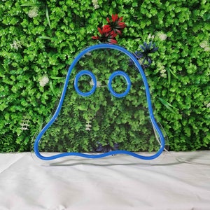 May include: A blue neon ghost sign with two round eyes. The sign is made of a flexible tube and is mounted on a white surface in front of a green wall with small white and red flowers.