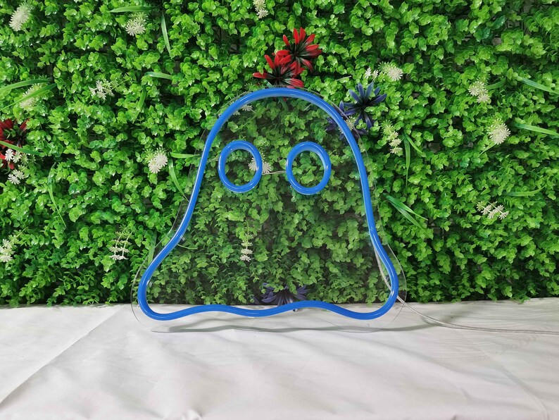 May include: A blue neon ghost sign with two round eyes. The sign is made of a flexible tube and is mounted on a white surface in front of a green wall with small white and red flowers.