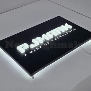 Könnte beinhalten: Ein rechteckiges schwarzes Schild mit dem weißen Text "PJMARK" und "A MARK OF EXCELLENCE". Die Buchstaben sind beleuchtet und das Schild hat einen weißen Lichtrand. Das Schild befindet sich auf einer weißen Oberfläche.