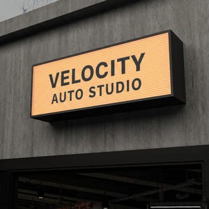 Może przedstawiać: Prostokątny szyld z napisem "VELOCITY AUTO STUDIO" w czarnych literach. Szyld ma jasno pomarańczowe tło i czarną ramę. Jest zamontowany na szarej betonowej ścianie.