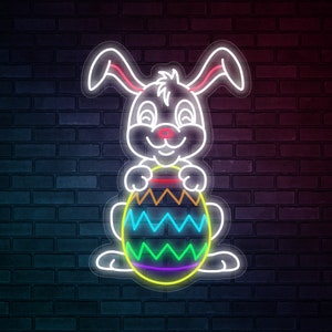 May include: Neon sign depicting a white Easter bunny with pink ear and nose accents, holding a colourful egg. The egg is decorated with zig-zag patterns in various colours. The sign is mounted on a dark brick wall.