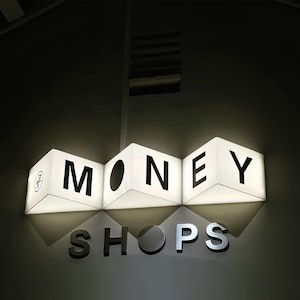 May include: Illuminated sign with the word "MONEY" in large, black capital letters. Below, the word "SHOPS" is displayed in silver letters. The sign is hanging from the ceiling.