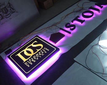 Puede incluir: Un letrero negro con letras doradas que dice "LOS Diplomatic Corporate Services" con luces LED moradas alrededor del perímetro. El letrero está sobre una superficie blanca con un letrero negro que dice "ISTOIA" con luces LED moradas.