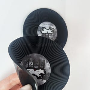 May include: Two black vinyl record-shaped coasters. The center of each coaster features a design with the number "4444" and a white graphic. The coasters have a ridged edge and are on a white background.
