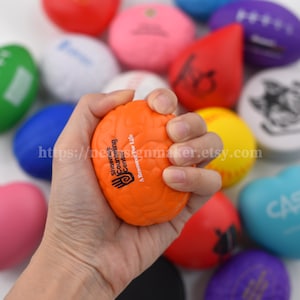 May include: An orange brain-shaped stress ball is held in a hand, with the text "Shawnee Counseling Center" and a logo. Other colorful stress balls, including footballs and spheres, are in the background. The Etsy shop URL is visible.