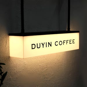 May include: Illuminated rectangular sign with the text "DUYIN COFFEE" in black capital letters. The sign is suspended from a black metal frame and emits a warm, inviting glow. The sign is mounted on a textured, off-white wall.